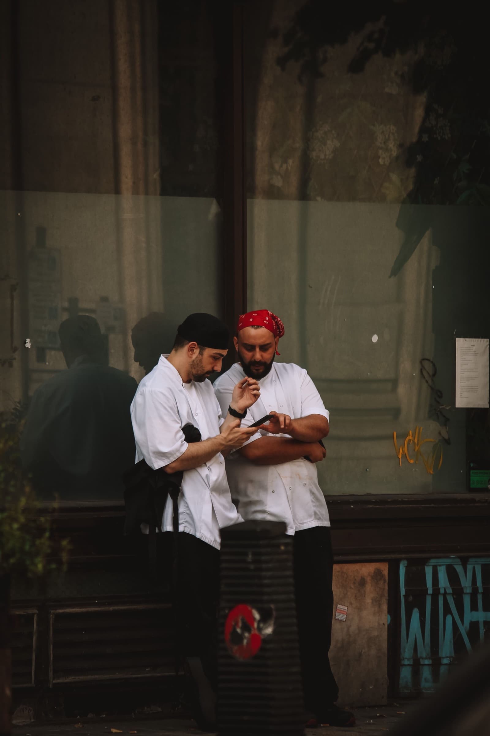 Two chefs in white jackets on a break outside a shopfront, looking at a phone together.