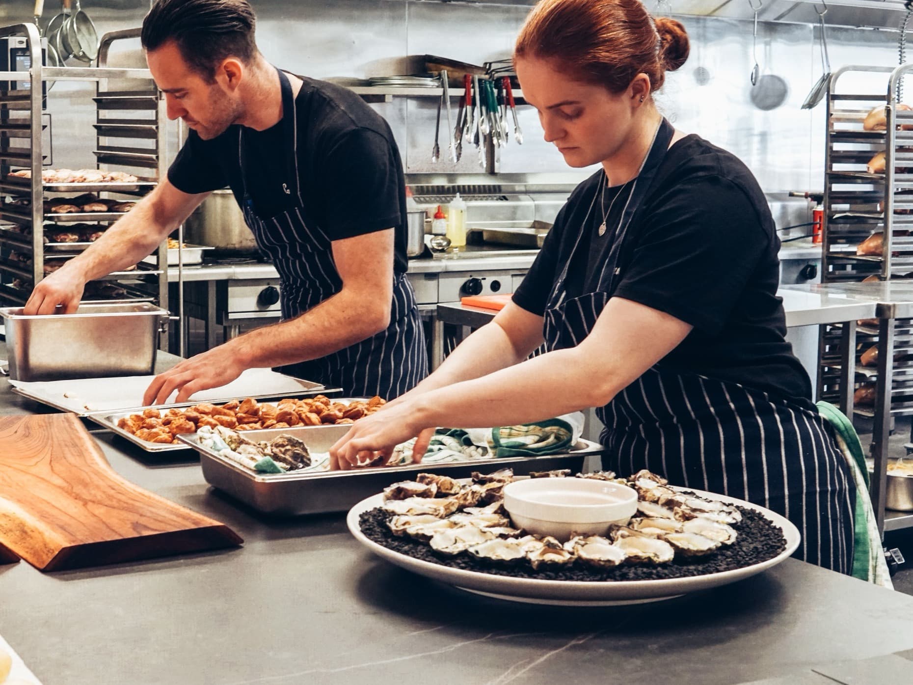 Two chefs in striped aprons preparing trays of food at a kitchen prep bench.